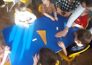 Dzieci z grupy II przy stoliku zwijają plaster wosku na świecę
