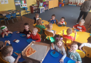 Dzieci z grupy II siedzą przy stolikach i jedzą pizzę
