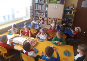 Dzieci z grupy II siedzą przy stolikach i jedzą pizzę
