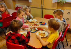 Dzieci z grupy V siedzą przy stoliku i dodają składniki do makaronu