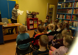 Dzieci siedzą na krzesłach i słuchają wiersza czytanego przez panią bibliotekarkę