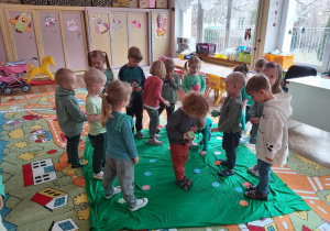 Dzieci z grupy I i III chodzą stoją na zielonej tkaninie i zbierają kwiatki