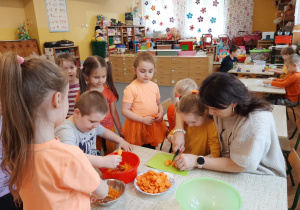 Pani dyrektor przy stoliku pomaga Marysi kroić marchewkę a dzieci patrzą