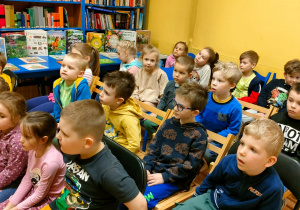 Dzieci siedzą na krzesłach i słuchają czytanego opowiadania