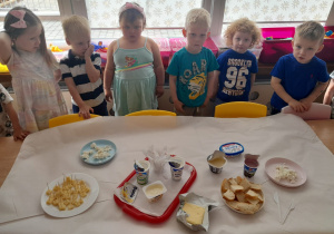 Kilkoro dzieci z grupy I stoi przy stole z produktami mlecznymi