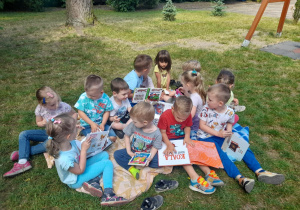 Dzieci z grupy III z książkami siedzą na kocu w ogrodzie