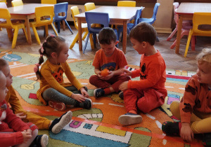 Dzieci siedząc na dywanie podają sobie dynie