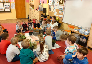 Dzieci z grupy Mrówki siedzą na dywanie i słuchają, jak Pani zielarka pokazuje zioła