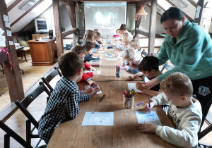 Dzieci przy stole kolorują szablon ozdoby świątecznej, pani Marta stoi i pomaga
