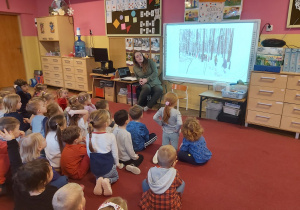 Pani leśnik siedzi przy tablicy, a dzieci oglądają slajd przedstawiający las