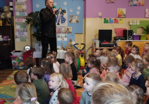 Pan Witek stoi i trzyma w ręce mikrofon, a dzieci siedzą na dywanie