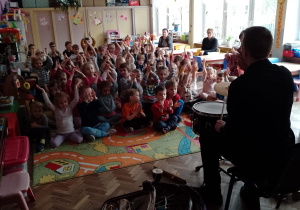 Dzieci siedząc podnoszą ręce do góry podczas, gdy pan Piotr gra na perkusji