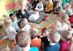 Pan Rafał kuca przed fantomem, a dzieci siedzą na dywanie