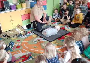 Pan Rafał kuca przed fantomem, dzieci siedzą na dywanie