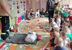 Pan Rafał stoi przed fantomem, pani Marta pokazuje kurtkę ratownika dzieciom, które siedzą na dywanie