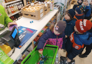 Dzieci stoją przy kasie w sklepie, maja wkłada do koszyka lizaki