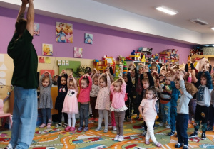 Dzieci naśladują prowadzącego i stojąc na palcach trzymają złączone ręce w górze