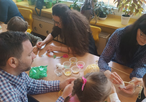 Dzieci z rodzicami przy stolikach ozdabiają jajka