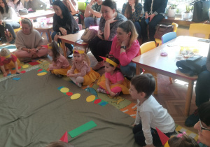Dzieci siedząc na dywanie układają na kartonikach sylwety pisanek, a rdzice siedzą przy stolikach