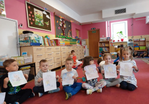 Kilkoro dzieci siedzi na dywanie i trzyma w rękach karty pracy