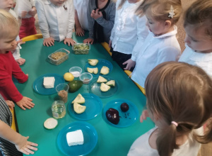 Dzieci z grupy Biedronki stoją przy stoliku, na którym leżą produkty do przygotowania śniadania