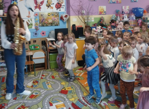 Pani muzyk gra na saksofonie, a dzieci poruszają się do muzyki