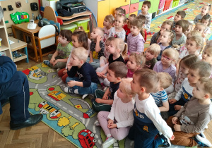 Dzieci siedzą na dywanie i słuchają strażnika miejskiego