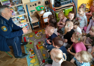 Dzieci siedzą na dywanie i słuchają strażnika miejskiego