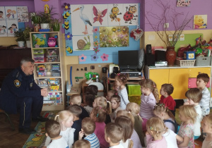 Dzieci siedzą na dywanie i słuchają strażnika miejskiego
