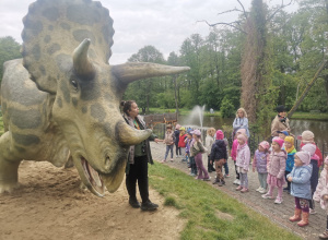 Pani przewodnik stoi przy dinozaurze, dzieci słuchają