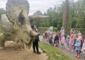 Pani przewodnik stoi przy dinozaurze i opowiada dzieciom o nim