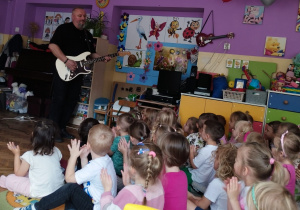 Gość gra na gitarze, a dzieci siedzą na dywanie