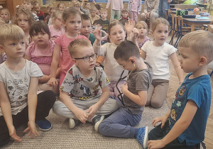 Dzieci siedza na dywanie podczas spotkania, a jeden chłopiec zakłada stetoskop