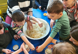 Dzieci z rodzicami przy stolikach ugniatają kapustę
