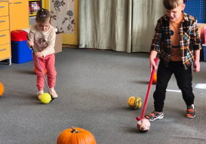 Dwoje dzieci toczy kijkiem piłkę między dyniami