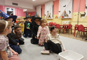 Dzieci siedzą w dwu rzędach przy wróżbie z wrzucaniem grosików do miski