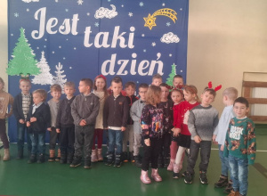 Dzieci stoją w sali gimnastycznej na tle dekoracji świątecznej