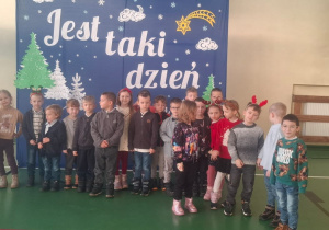 Dzieci stoją w sali gimnastycznej na tle dekoracji świątecznej