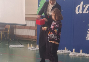 Dziewczynka odczytuje tekst na karteczce, a nauczycielka stoi obok i trzyma pudełko