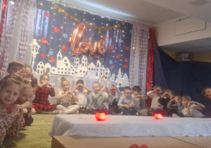Dzieci siedza na scenie z rączkami ułożonymi w kształcie serca