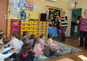 Dzieci siedzą na dywanie i słuchają piosenki śpiewanej przez dwiepanie prowadzące