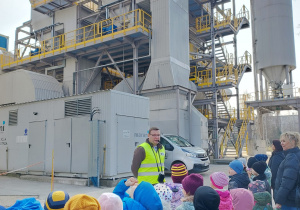 Dzieci stoją przed budynkiem elektrociepłowni i słuchają pracownika
