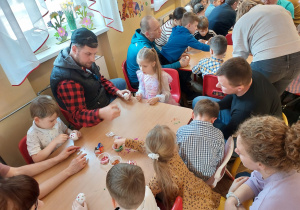 Dzieci z rodzicami przy stolikach ozdabiają jajka