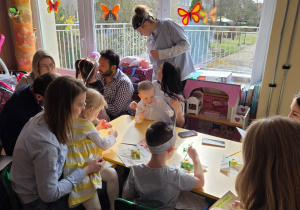 Dzieci z rodzicami przy stolikach malują zajączki