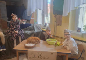 Dwoje dzieci siedzi na scenie, a dwoje przy stole podczas przedstawienia