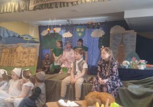 Dwoje dzieci siedzi na scenie, troje stoi, a kikoro pod sceną podczas przedstawienia