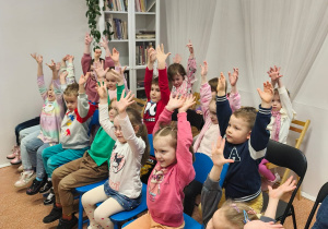 Dzieci siedzą w czytelni i podnoszą ręce w górę