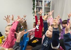 Dzieci siedzą w czytelni i podnoszą ręce w górę