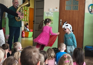 Chłopiec w czapce byka goni dziewczynke z chustką, a muzyk gra na instrumencie