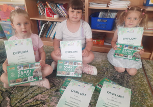 Trzy dziewczynki siedzą na dywanie z dyplomami i upominkami